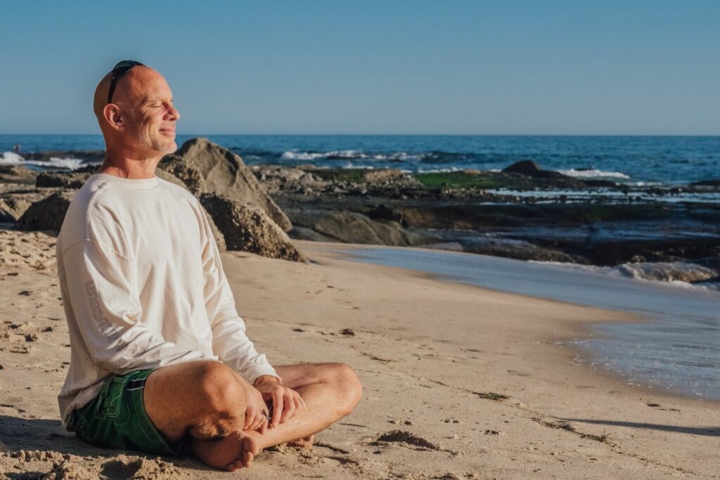 Benefits of mindfulness - Man sitting cross-legged smiling with eyes closed on beach